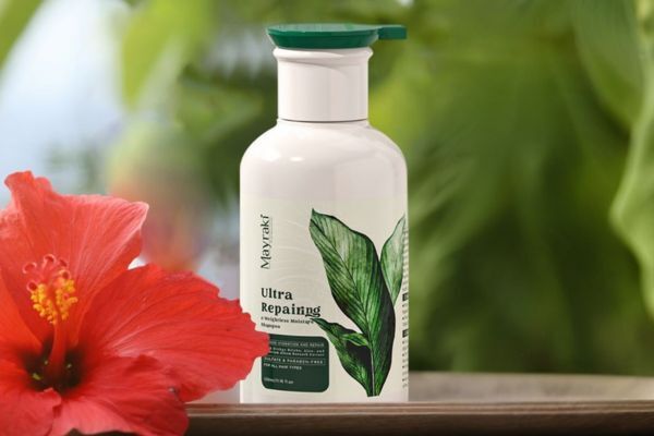 A bottle of Mayraki Ultra Repairing & Weightless Moisture Shampoo stands on a wooden surface with a vibrant red hibiscus flower in the foreground and lush green foliage in the background. The packaging features botanical artwork, emphasizing its hydrating, natural formula for hair repair.