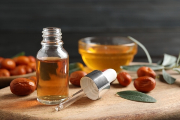 Close-up of a glass dropper bottle filled with golden oil, surrounded by jojoba seeds, a bowl of oil, and sage leaves on a wooden surface, representing the natural ingredients used by Mayraki to help fix a patchy beard.