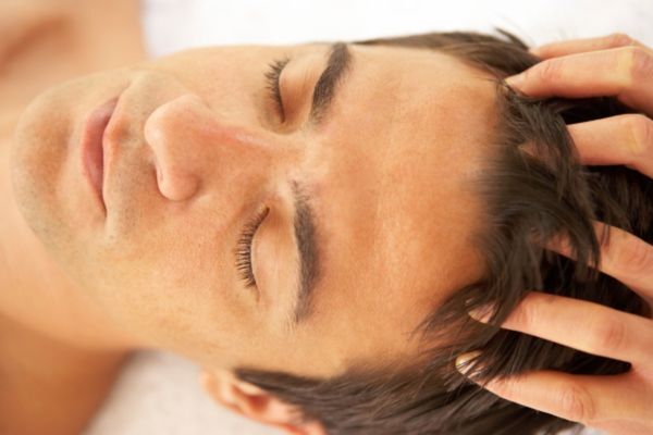 Natural and Gentle Prevention Methods A man with closed eyes lies on his back while receiving a relaxing scalp massage. The image conveys a sense of calm and rejuvenation, complementing Hair Mayraki’s approach to scalp care as part of a soothing self-care routine.
