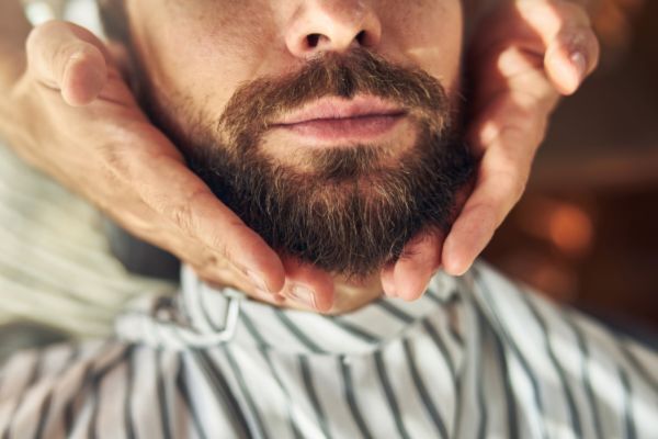 A close-up of a man’s well-groomed beard being gently framed by a barber’s hands. He’s wearing a striped cape, suggesting he’s in a professional grooming session.