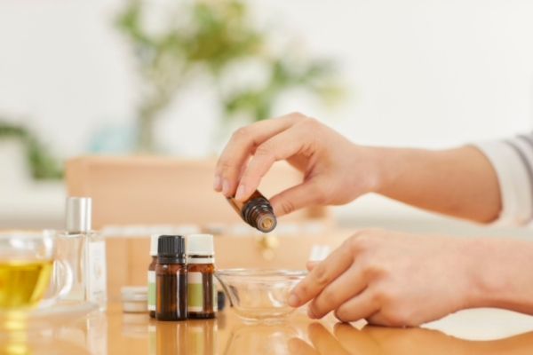Natural Ingredients and Essential Oils for Beard Growth Person's hand pouring oil from a small amber bottle into a glass dish on a wooden table, surrounded by various essential oil bottles and grooming products.