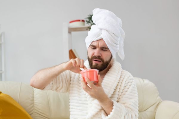 A bearded man in a bathrobe and towel turban sits on a couch while examining a jar of cream with a puzzled expression, representing a search for natural remedies to treat beard dandruff (beardruff).