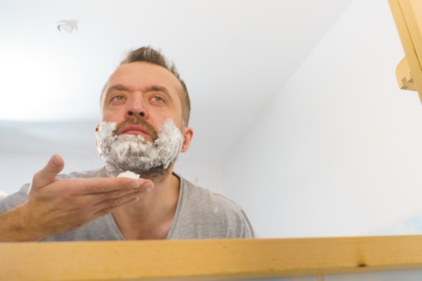 Man applying shaving cream in front of a bathroom mirror, preparing his beard for grooming—an essential step in maintaining beard health and protection, especially before outdoor exposure.