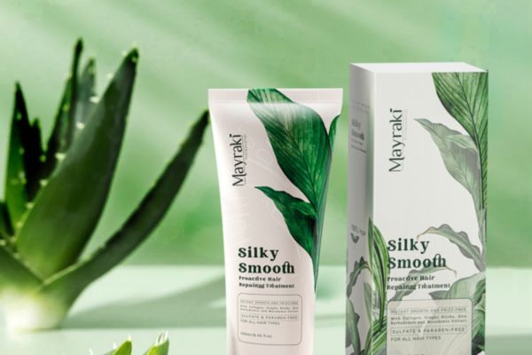A tube and box of Hair Mayraki’s Silky Smooth Proactive Hair Repair Treatment are displayed on a green background with aloe leaves in soft focus. The natural setting highlights the product’s botanical ingredients and its commitment to repairing and smoothing all hair types.