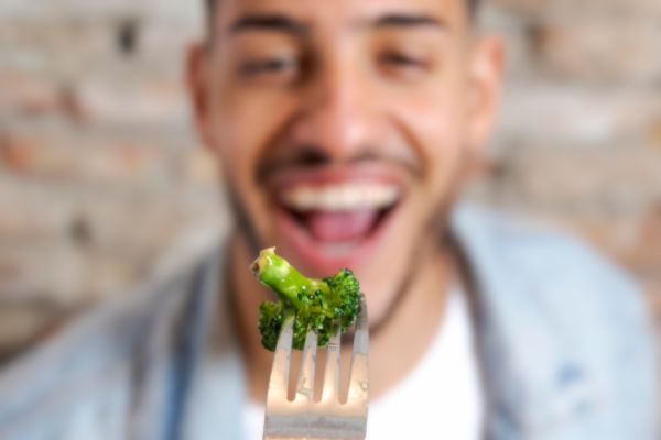 Close-up of a man smiling with an open mouth while holding a fork with a single piece of broccoli in focus. The playful image emphasizes healthy eating or a joyful mealtime moment.