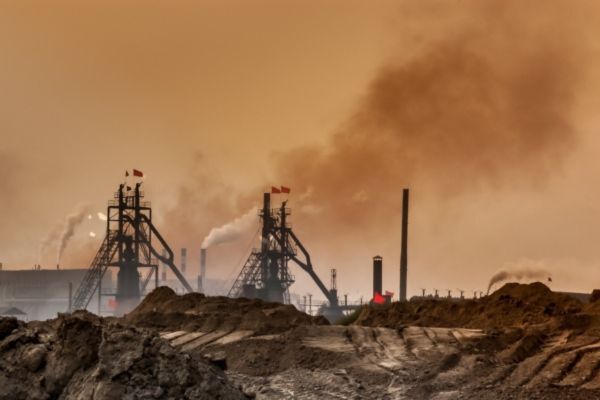 Industrial scene with smokestacks emitting thick pollution into a hazy, orange-tinted sky, surrounded by piles of dirt and barren land. The image emphasizes environmental degradation and air pollution caused by heavy industry.