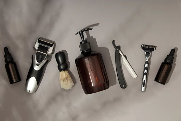 Flat lay of men's grooming tools including two small dropper bottles, an electric shaver, a shaving brush, a brown pump bottle, a straight razor, and a manual razor arranged neatly on a neutral background. The setup highlights essential items for beard and facial grooming routines.
