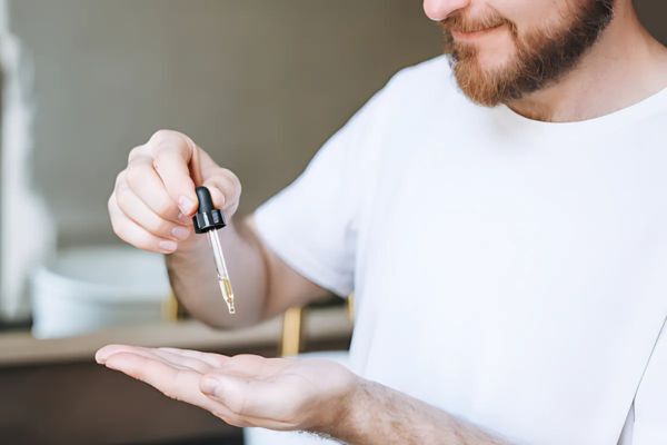 Smiling bearded man in a white shirt uses a dropper to apply beard oil into his palm, preparing for grooming. The scene suggests a personal care routine focused on beard maintenance.