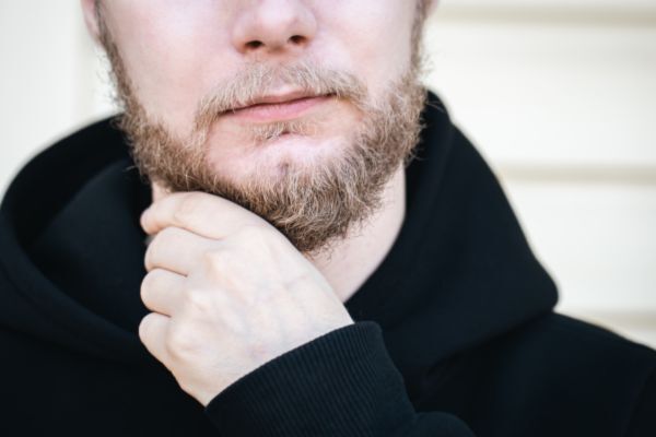 A close-up of a man with a patchy, light brown beard and fair skin, gently holding his chin. He is wearing a black hoodie, and the image highlights uneven beard growth and possible early signs of graying.