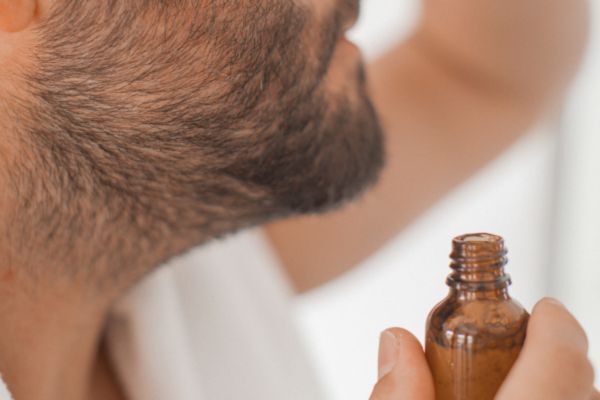 Close-up of a bearded man holding an open bottle of beard oil near his chin, preparing for Mayraki CopperGro Pro Beard Oil usage as part of a grooming routine focused on hydration and growth.