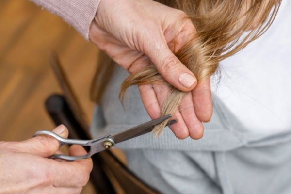 Close-up of a person cutting the ends of long, light brown hair with a pair of silver scissors. The hands are steady and focused, emphasizing a precise at-home hair trim