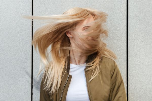 A woman with long, blonde, silky hair flipping her head, causing her hair to fan out around her face, wearing a white shirt and olive green jacket against a textured light gray wall.
