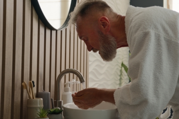 Older man in a white robe washing his face at a bathroom sink, a key step in softening facial hair and reducing beard tangles as part of a proper grooming routine.