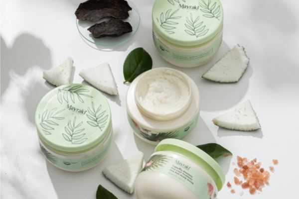 A flat lay of Hair Mayraki scalp scrub jars surrounded by natural ingredients like coconut pieces, tea leaves, and pink Himalayan salt. The clean, sunlit setup emphasizes Hair Mayraki’s use of natural exfoliants in promoting scalp health and reducing dandruff.