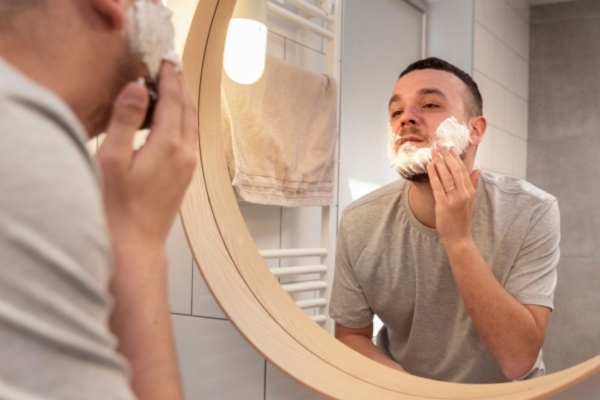 A man stands in front of a round bathroom mirror applying shaving cream to his face, mid-morning routine in a softly lit, modern bathroom.