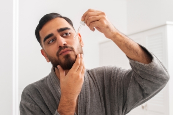 A man in a gray robe applies beard oil with a dropper while standing in a bright, minimal bathroom. His focused expression and grooming routine highlight self-care and beard maintenance.