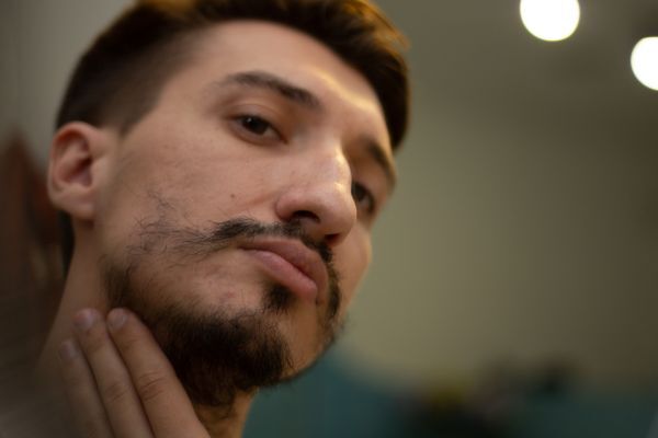 A man closely examines his patchy beard growth in the mirror, gently touching his jawline with his fingers. His expression is serious, highlighting concern or curiosity about his facial hair or skin condition.