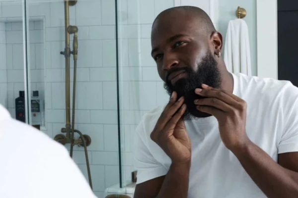 A man examining his full beard in the mirror with both hands, checking for split ends and uneven growth, visually representing the Signs It’s Time to Trim Your Beard.