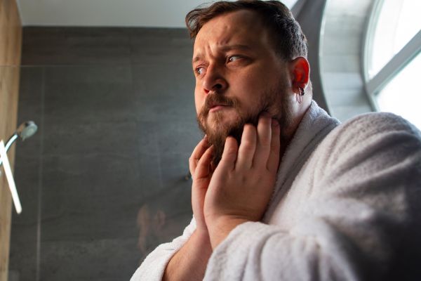 A man in a white bathrobe stands in front of a bathroom mirror, gently touching his beard with a concerned expression. The image suggests he may be noticing irritation or damage, possibly linked to environmental stress or climate-related skin and hair issues.