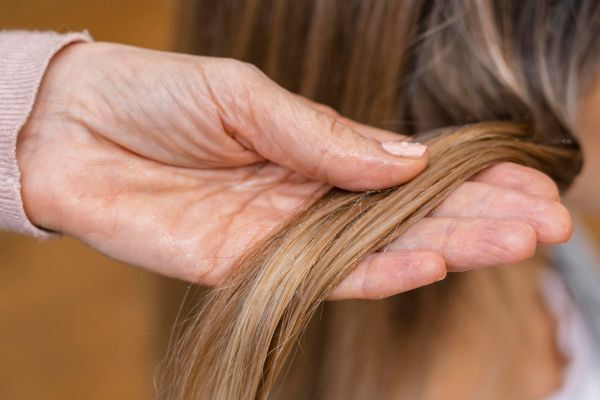 A close-up of a person gently holding a section of light brown hair between both hands, showing dry, slightly frizzy strands. The image suggests assessing hair health and may indicate the need for deep conditioning or a protein treatment.