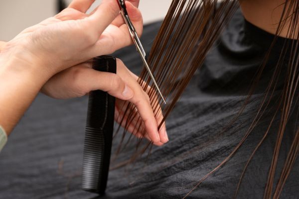 A hairstylist trims the ends of straight, damp hair using scissors and a comb, focusing on precision to prevent split ends. The close-up captures a salon setting and highlights the importance of regular haircuts for maintaining healthy hair.