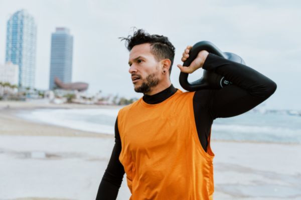 Man wearing a bright orange sleeveless workout shirt over a black base layer carries a kettlebell over his shoulder while standing on a beach, with city buildings and ocean waves in the background, representing an active lifestyle.