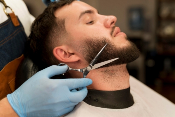 A man reclines in a barber chair as a gloved barber carefully trims his full beard with scissors, focusing on the neckline for a clean edge.
