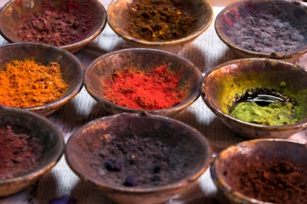 Nine small metal bowls filled with brightly colored powdered pigments, including red, orange, green, and purple, arranged on a newspaper-covered surface.