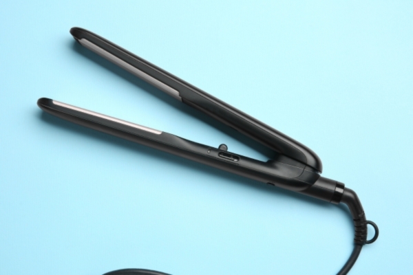 Black flat iron hair straightener with silver plates resting open on a solid light blue background, showing its sleek design and power switch.