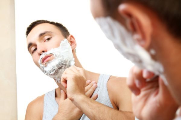 A man in a gray tank top looks into the mirror as he carefully shaves his face with shaving cream applied. His focused expression shows he’s concentrating on maintaining clean, precise lines.
