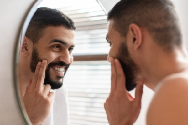 Bearded man smiling confidently while pointing at his cheek in the mirror, suggesting satisfaction with his skincare or grooming routine.