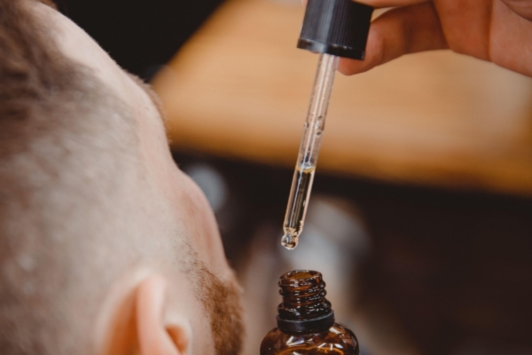 Close-up of a man’s face as a dropper releases beard oil from an amber bottle, with the focus on the droplet about to fall. The scene highlights personal grooming and beard care application.