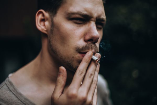 A man smokes a cigarette with a serious expression, holding it close to his lips as smoke curls upward. The background is blurred, drawing attention to his face and the act of smoking.
