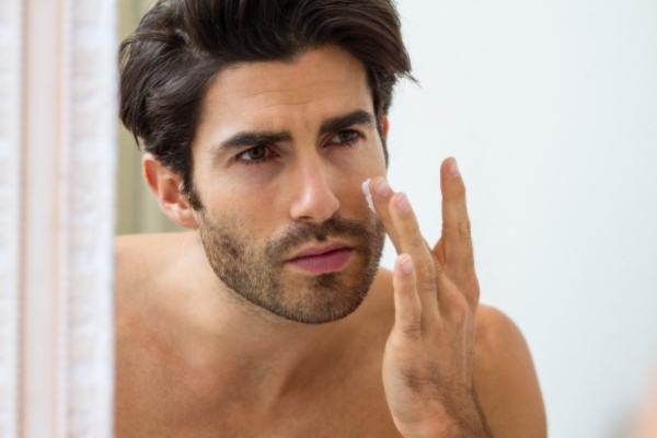 Close-up of a shirtless man with short dark hair applying face cream to his cheek while looking in the mirror, with a focused expression.