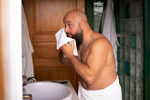 Bald man with a full beard stands at a bathroom sink, patting his face dry with a towel while wearing a white towel wrapped around his waist. The image suggests a grooming or skincare routine in a home setting.