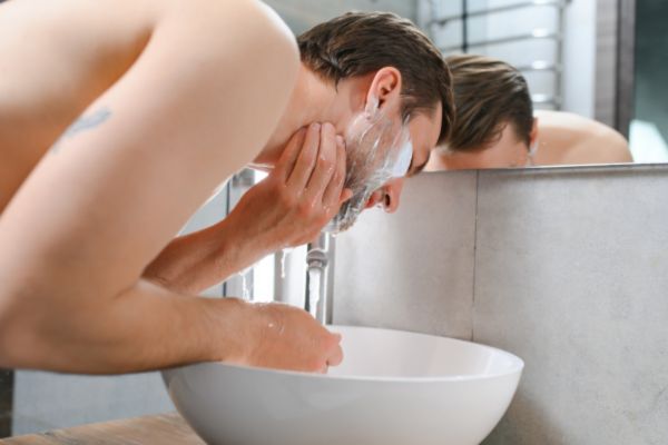 Shirtless man washing his face over a modern white basin, applying cleanser to his skin in front of a mirror, suggesting part of a daily grooming routine.