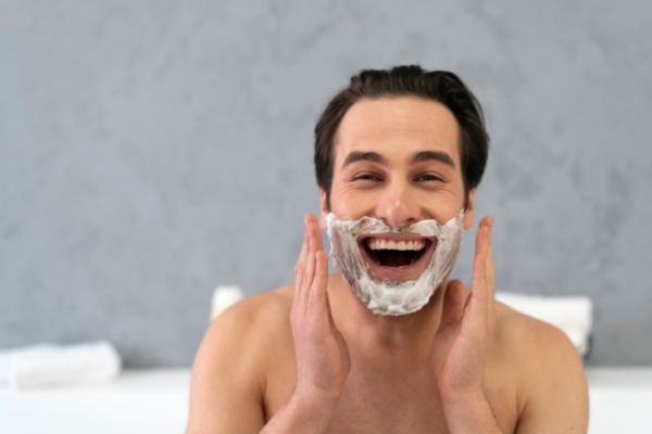 Smiling man with shaving cream on his face applies it in front of a gray background, giving a cheerful and relatable moment during a grooming routine.