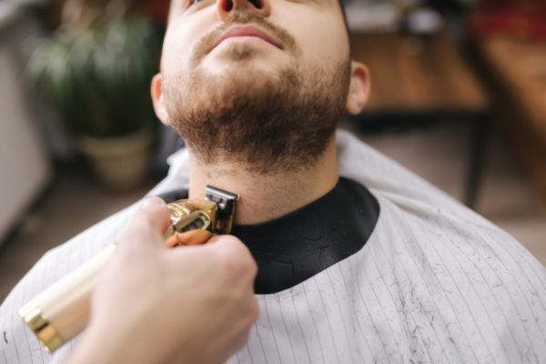 A barber trims a man's beard with electric clippers while he reclines in a chair wearing a striped barber cape, representing precision grooming and styling techniques.