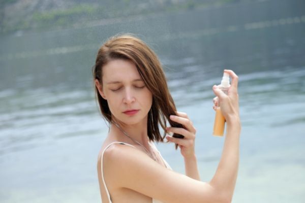 Woman standing by a calm body of water, eyes closed as she sprays a protective mist onto her hair. Dressed in a white tank top, she demonstrates summer hair care and UV protection in a sunny, outdoor setting.
