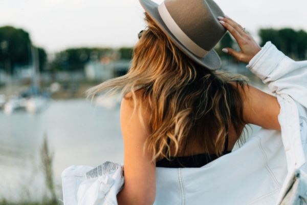 Woman with sun-kissed wavy hair facing a waterfront, wearing a tan hat and white jacket draped over her shoulders. The image captures the effect of sun exposure on hair while highlighting stylish sun protection.