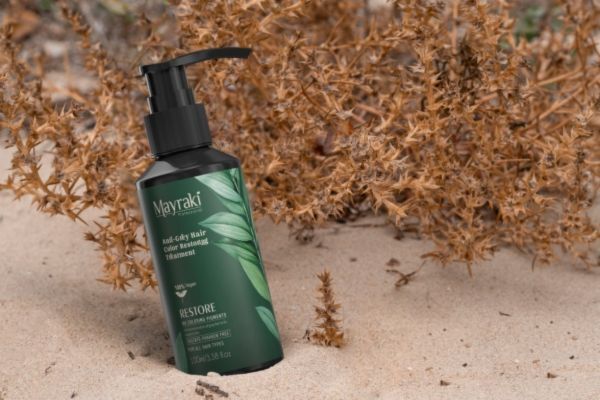 This image features a bottle of Mayraki's Anti-Gray Hair Color Restoring Treatment resting on sandy ground in front of dried, brown desert plants. The green packaging highlights natural ingredients and promises to restore hair color without harsh chemicals, emphasizing eco-conscious beauty.