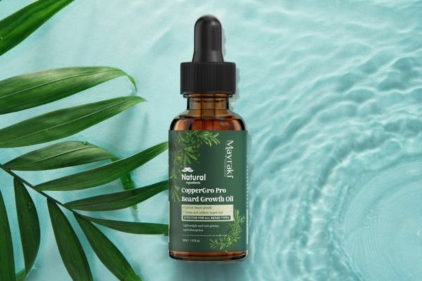 A bottle of Mayraki CopperGro Pro Beard Growth Oil placed beside tropical green leaves on a rippling aqua surface, evoking a clean, natural aesthetic. The amber glass bottle with a green label emphasizes its organic formula for beard strength and growth.