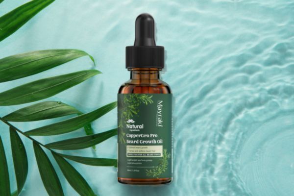 A dropper bottle of Mayraki CopperGro Pro Beard Growth Oil placed on a textured water background with green tropical leaves, labeled as natural and designed to enhance beard thickness and strength.