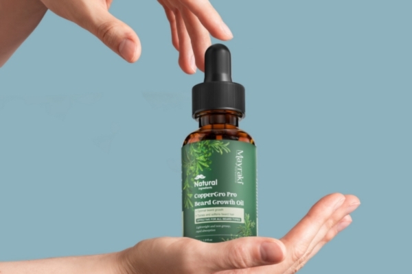 Hands carefully frame a bottle of Mayraki CopperGro Pro Beard Growth Oil against a light blue background, showcasing the green label with natural ingredients and dropper cap.