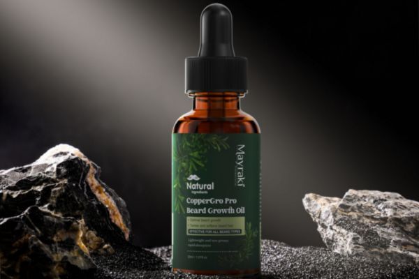 A bottle of Mayraki Natural CopperGro Pro Beard Growth Oil stands upright between dark textured rocks under dramatic lighting. The amber glass bottle features a green label with leafy graphics, emphasizing its natural, plant-based formula for beard care.