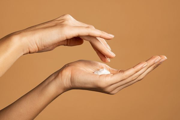 Close-up of a person dispensing a small amount of white cream or lotion onto their palm against a warm beige background. The image emphasizes proper product usage or the right amount of application for skincare or haircare routines.