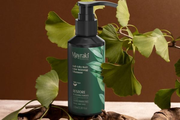 A bottle of Hair Mayraki’s Anti-Gray Hair Color Restoring Treatment is displayed upright against a backdrop of ginkgo leaves and a neutral-toned surface. The elegant dark green label and natural elements emphasize the brand’s plant-based, restorative hair care philosophy.