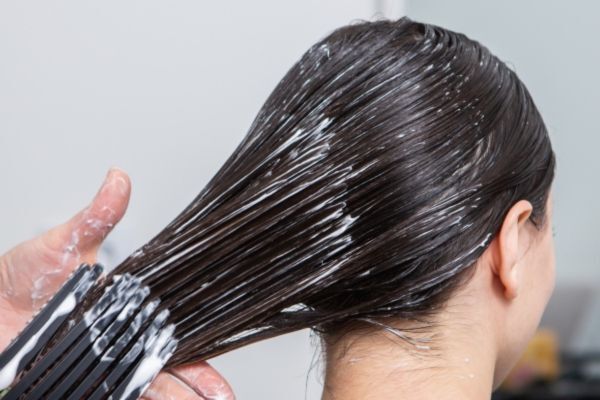 A close-up of a person with dark, wet hair having a keratin treatment applied. A hand is seen using a wide-tooth comb to evenly distribute a creamy, white keratin product through the hair, creating a sleek, glossy appearance.