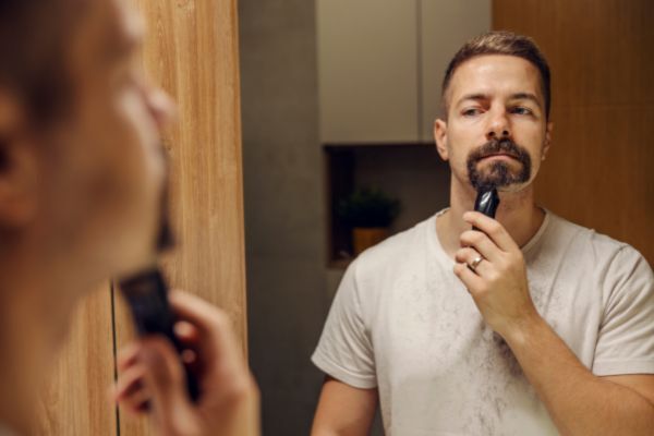 A man looks into the mirror while using an electric trimmer to shape his beard. He’s wearing a white t-shirt with visible beard trimmings, indicating a mid-routine grooming moment.
