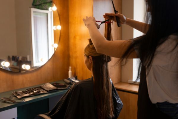 A hairstylist uses scissors to trim the long, straight hair of a seated woman in a salon. The room is warmly lit with a round mirror and hair tools neatly arranged on the counter.